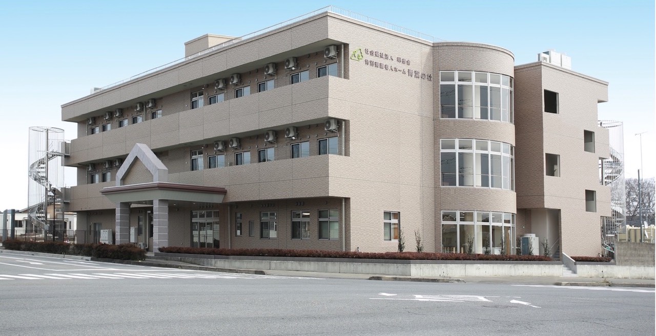 3階建てのベージュ色介護施設。玄関・駐車場・外構が見える。